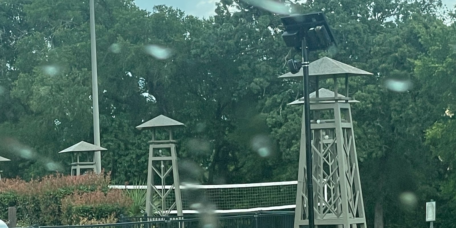 Seen through a rain-speckled car window, license plate reader cameras mounted on a wooden tower beside a fenced pool area